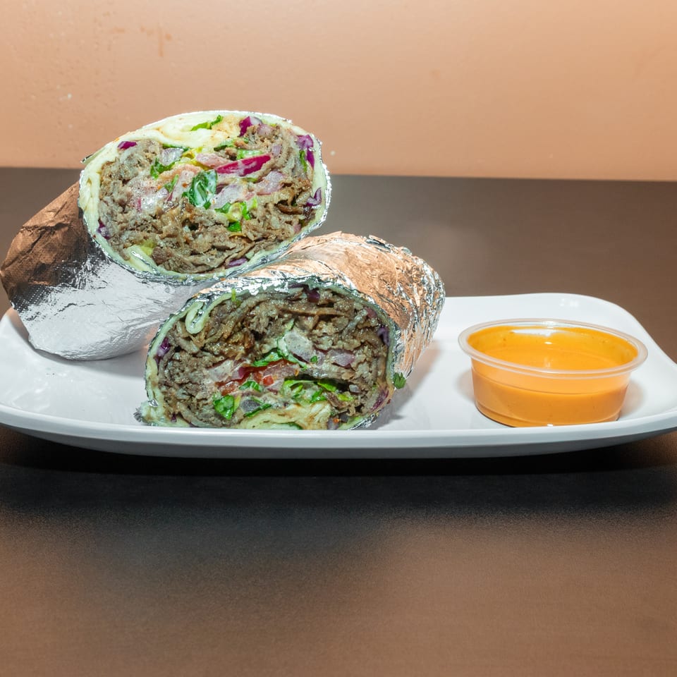 Two foil-wrapped meat shawarma halves with lettuce and vegetables sit on a white plate beside orange dipping sauce.