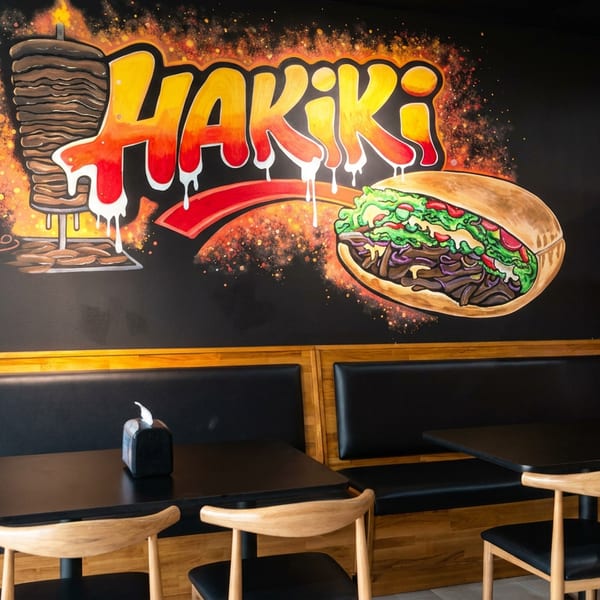 Black-walled restaurant dining area with booth seating, wooden chairs, tables, Hakiki sandwich mural, and Coca-Cola refrigerator.