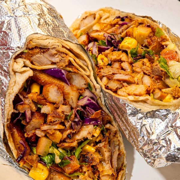 Two foil-wrapped shawarma wraps filled with sliced meat, corn, cabbage, lettuce, and colorful vegetables on a white plate.