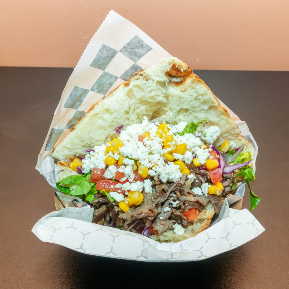 Loaded pita sandwich filled with sliced meat, lettuce, tomatoes, corn, red onion, and crumbled white cheese.