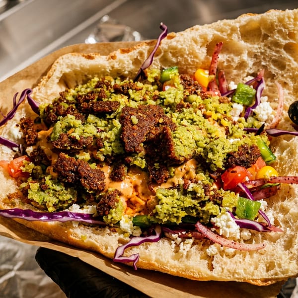 Loaded flatbread sandwich with falafel, green herbs, vegetables, purple cabbage, and creamy sauce, held in gloved hands.