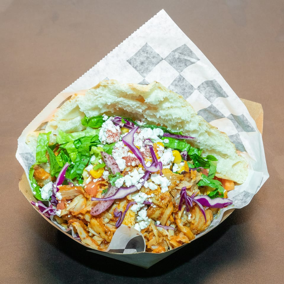 Loaded Greek-style pita topped with chicken, lettuce, tomatoes, corn, red cabbage, onions, and crumbled feta in a paper-lined tray.