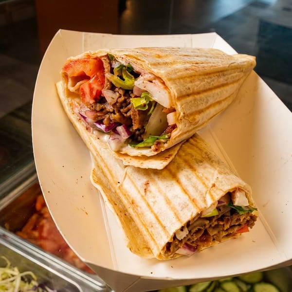 Two grilled wraps filled with seasoned meat, vegetables, and sauce sit in a paper tray above a restaurant ingredient counter.